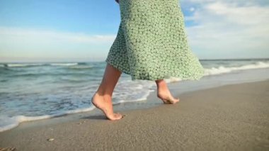 Yakından çıplak ayaklı yeşil elbiseli, çekici, beyaz tenli genç bir kadın sabahın erken saatlerinde deniz kenarında ıssız bir plaj boyunca geziniyor. Yavaş çekim.