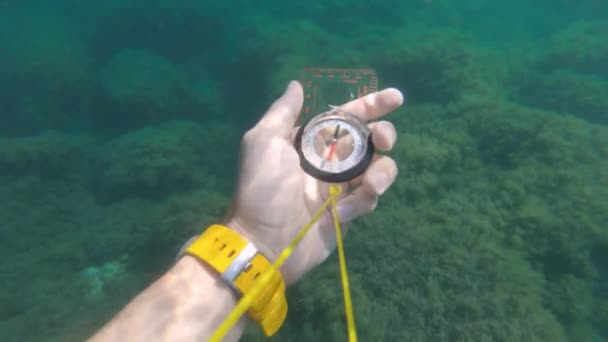 Första person syn på en manlig hand som håller en plast magnetisk kompass roterar från sidan och söker rätt riktning under vattnet. — Stockvideo