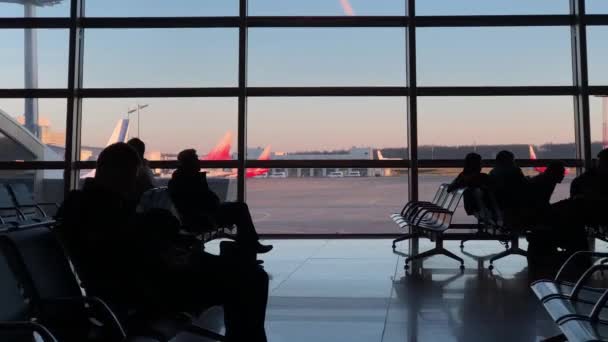 Silhouettes de personnes assises dans le salon de l'aéroport avant le départ. Des avions se tiennent derrière la vitre. mouvement de caméra panoramique — Video