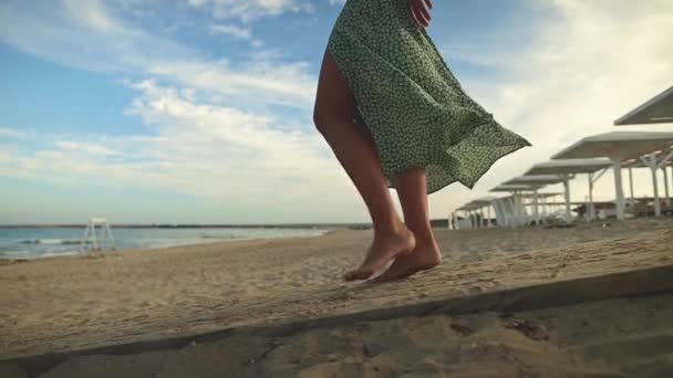 Piernas de una joven en cámara lenta caminando descalza por la playa. Dejando huellas en la arena Turista de vacaciones junto al mar. Mujer en un hermoso vestido revoloteando en el viento — Vídeo de stock