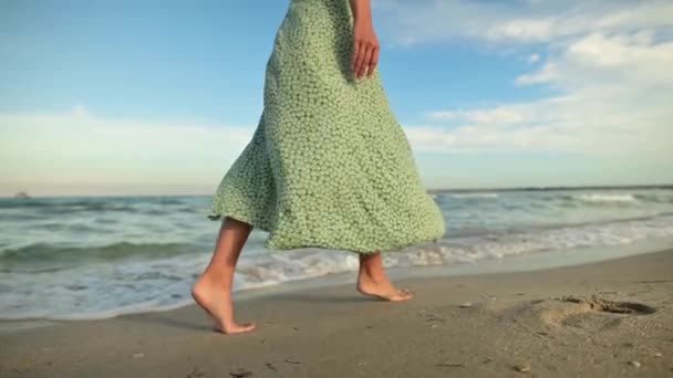 Atractivas piernas delgadas de una mujer en cámara lenta caminando descalza por la playa por la mañana temprano. Turista en un vestido verde claro en una playa dorada — Vídeo de stock
