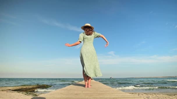 Une jolie jeune femme caucasienne dans une robe verte d'été et un chapeau de paille se tient sur une jetée en bois sur le bord de la mer et danse — Video