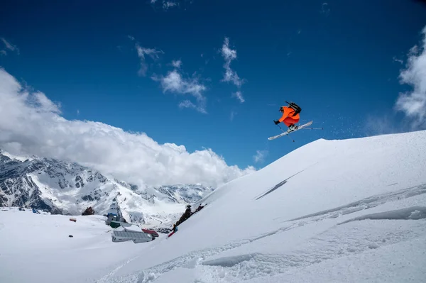 Atleta professionista giovane sciatore maschile in tuta arancione vola sopra le montagne dopo essere saltato da sporgenze innevate. Comunità sportiva freeride sullo sfondo di cime innevate — Foto Stock