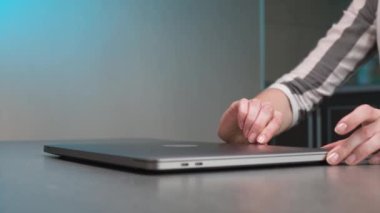 Genç beyaz bir kadının elleri mavi bir arka ışığa karşı gri bir dizüstü bilgisayar açıyor. Sığ alan derinliği