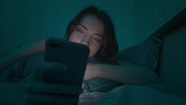 Genç beyaz kadın, alacakaranlıkta yatmadan önce akıllı telefonunu kullanıyor. Mobil bağımlılık kavramı mavi ışık gözler için zararlıdır. — Stok video