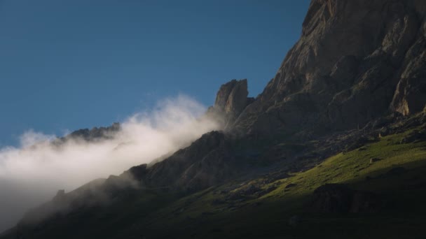 Verano video paisaje baja cubierta de nubes fluye alrededor de acantilados épicos en una noche de verano al atardecer. Prados verdes con hierba al pie de las montañas rocosas — Vídeo de stock