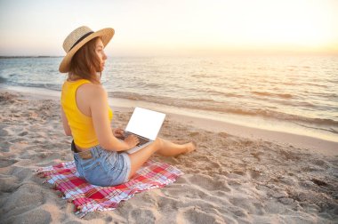 Genç beyaz şortlu ve kumsalda şapkalı bir kadın, kumsalda oturuyor, yazın bilgisayardaki sosyal ağları tarıyor denizin arka planına karşı..