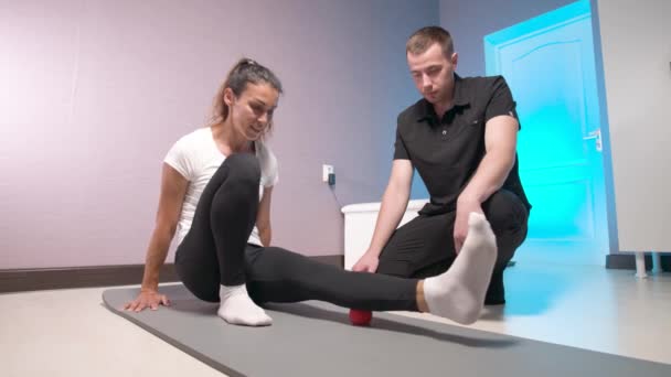Młody biały mężczyzna masażysta pokazując i kontrolując masaż mięśnia sercowego powięzi nóg do kobiety klienta. Trening samomasażu przy użyciu piłki do masażu — Wideo stockowe