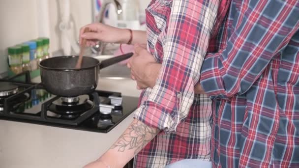 Young happy husband hugs and kisses smiling wife while standing by the stove in the kitchen, affectionate couple prepares healthy food. Home life and everyday life of a young family — Stock Video