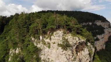 Uzun saçlı, sıska, siyah tişörtlü ve şortlu bir adam uçurumun kenarında duruyor ve etrafındaki orman ve dağlara bakıyor. Hava görünümü
