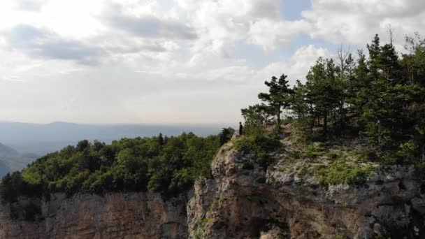 A young man in a black T-shirt and shorts stands on the edge of a cliff in front of a cliff against the backdrop of a forest mountain valley in summer. Aerial view — Stock Video