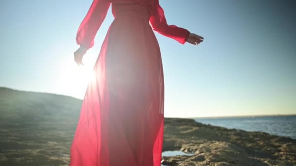 Mujer caucásica joven en cámara lenta en un vestido rojo aleteo suelto está caminando a lo largo de la costa rocosa. Profundidad de campo superficial. Concentración compensatoria. — Vídeo de stock