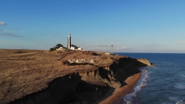 Vista aérea de un faro solitario rodeado de edificios técnicos se encuentra en un promontorio rocoso desierto en la orilla del mar. Oceánica costa de la tarde. Faro al atardecer. Bajo perfil. grano de película — Vídeo de stock