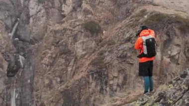 Elinde kamera olan genç bir erkek fotoğrafçının hava görüntüsü bir vadideki yüksek bir kayanın üzerinde duruyor ve baharda kar fırtınası ve kar yağışı sırasında bir alp şelalesinin fotoğraflarını çekiyor..