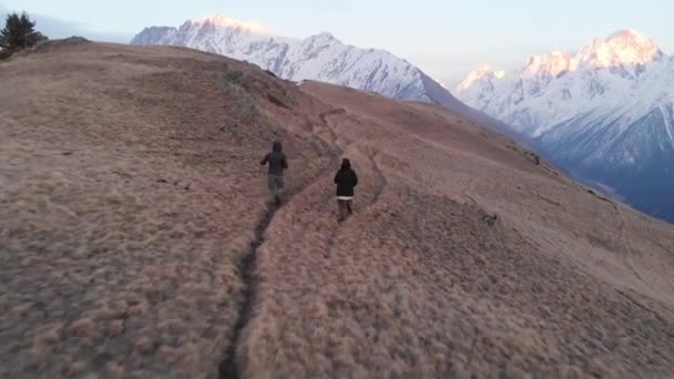 Una pareja amorosa de escaladores corre a lo largo de un sendero en una ladera de montaña en la tarde contra el telón de fondo de montañas cubiertas de nieve. — Vídeos de Stock