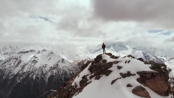 Ένας άνδρας ταξιδιώτης με σακίδιο στέκεται στην κορυφή ενός γκρεμού με φόντο χιονισμένες βουνοκορφές και χαμηλά σύννεφα. Αεροφωτογραφία. Πετώντας την κάμερα μπροστά από έναν άνθρωπο σε ένα βράχο storon — Αρχείο Βίντεο