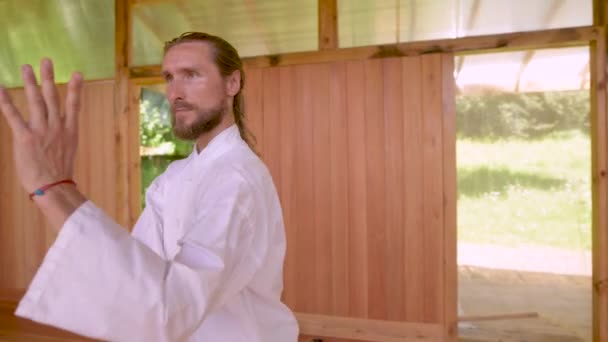Homem caucasiano barbudo em roupas leves para praticar está praticando qigong na sala de madeira do salão. Tradições modernas da prática energética — Vídeo de Stock