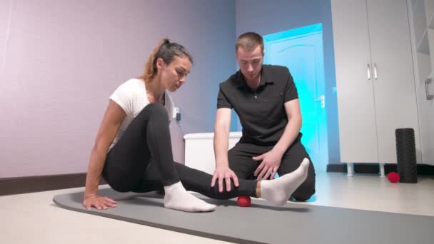 Молодий кавказький чоловічий масажист, який демонструє і контролює міофасциальний масаж фасції ніг до жіночого клієнта. Навчання самомасажу за допомогою масажного м'яча — стокове відео
