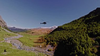 Dağlardaki gri bir helikopterin hava görüntüsü. Yeşil dağlarda teknik ulaşım uçuşları gerçekleştiriyor. Dağlarda kargo taşımacılığı. Sivil Havacılık