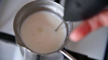 Gaz sobasında süt bulunan metal bir Türk kahve makinesinin yakın çekim görüntüsü. Bir kadının eli, elle tutulan bir elektrikli çırpı kullanarak köpüklü bir kıvama ulaşana kadar sütü köpürür.