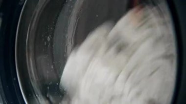Otomatik bir çamaşır makinesi kirli beyaz giysileri yıkar. Çamaşır makinesinde çamaşır yıkamak. Bir çamaşır makinesinin dönen davulunun yakın çekim videosu