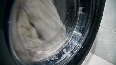 Bir zamanlar çamaşır makinesinde yıkanan, eski, sarı gri çamaşırların yakın çekimi. Deterjan köpükleri camdan aşağı damlıyor.