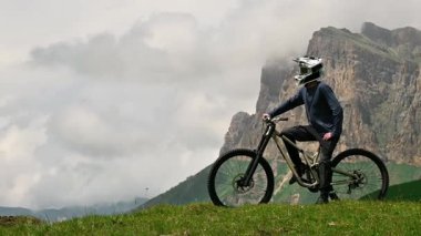İnsansız hava aracı dik çimen tepesinde dağ bisikletinin yanında duran atletik adamın yanından uçuyor. Dağların güzel manzarası. Doğada yapayalnız, hayatı yansıtan.
