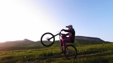 Miğferli ve maskeli genç bir sporcu gün batımında arka tekerlere binip pedal çeviriyor. Fpv insansız hava aracında hava görüntüsü
