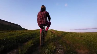 Miğferli ve maskeli genç bir sporcu gün batımında arka tekerlere binip pedal çeviriyor. Fpv insansız hava aracında hava görüntüsü.