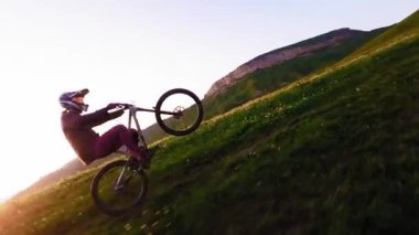 Miğferli ve maskeli genç bir sporcu gün batımında arka tekerlere binip pedal çeviriyor. Fpv insansız hava aracında hava görüntüsü