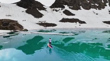 Şortlu bir adam kürek tahtasında durur ve gölde kürek çeker. Arka planda karlı dağlar ile yaz yürüyüşü sezonu başladı..