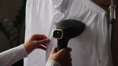 Bir kadın eli, buharlı bir giysi buharı kullanır, gömleğin üzerindeki kırışıklıkları düzeltir, sıcak bir buhar yayar. Evde buharlı demir.