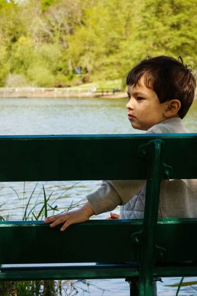 Liten pojke sitter på en bänk bredvid en sjö — Stockfoto