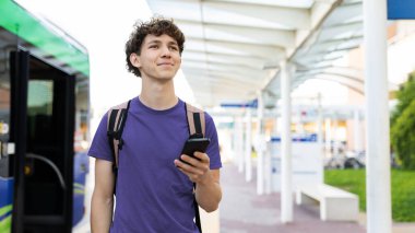 Young Caucasian man with backpack holding smartphone at airport terminal. Concept of travel, waiting, modern lifestyle, and exploring new destinations