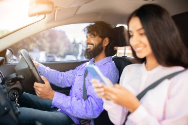 Hintli çift birlikte araba kullanıyor, erkek yola odaklanmış, kadın akıllı telefon kullanıyor. Modern yaşam tarzını, teknolojik alışkanlıkları ve ortak banliyö ve yakın mesafede bağlantıyı gösterir