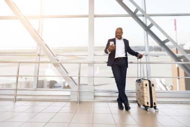 Havaalanı terminalinde korkuluklara dayanan valiziyle rahat bir Afrikalı Amerikalı işadamı, akıllı telefon, yanında bavul, iş seyahatinin keyfini çıkarırken gülümsüyor.
