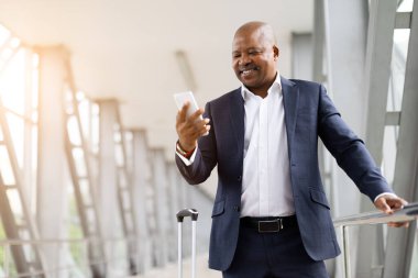 Kendinden emin Afrikalı Amerikalı işadamı havaalanı koridorunda akıllı telefondan mesajlar okurken gülümsüyor, uçuştan önce rahat ve mutlu, iş seyahati yaşam tarzı konsepti