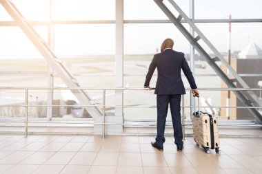 Havaalanı penceresinde elinde bavulla dikilen Afro-Amerikan işadamının dikiz görüntüsü, uçuştan önce uçakları izlemek, sakin ve profesyonel görünüm