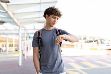 Sırt çantalı genç bir adam havaalanında beklerken kol saatine bakıyor. Seyahat çizelgesi, gecikme veya zaman yönetimi kavramı, kopyalama alanı