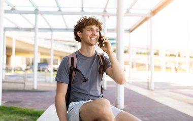 Sırt çantalı mutlu genç gezgin havaalanında akıllı telefonla konuşuyor. Neşeli sohbet ve pozitif seyahat duyguları parlak güneş ışığında, boşluğu kopyala