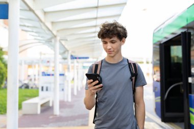 Havaalanı otobüs durağında duran sırt çantalı genç adam, ulaşımı beklerken akıllı telefonunu kontrol ediyor. Parlak gün ışığı ve gündelik seyahat ortamı