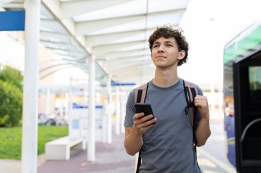 Sırt çantalı genç gezgin havaalanı yakınındaki otobüs durağında akıllı telefonunu tutarak yürüyor. Parlak sabah ışığı ve iyimser seyahat havası
