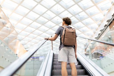 Sırt çantalı gezgin yürüyen merdivende duruyor, tırabzanları tutuyor ve yukarı bakıyor. Parlak tavan ve modern seyahat atmosferi, boşluğu kopyala