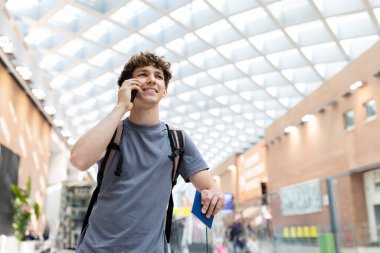Gezgin akıllı telefondan konuşuyor ve havaalanı terminalinde pasaportu tutuyor. Uçuş öncesi cep telefonuyla iletişim kuran neşeli genç adam, boşluğu kopyala