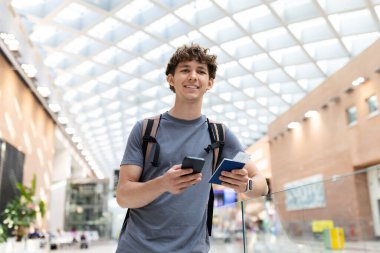 Havaalanında elinde akıllı telefon ve pasaportla gülümseyen genç bir adam. Heyecanlı yolcu giriş veya uçuş için hazır, terminalde duruyor, kopyalama alanı