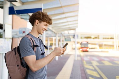 Havaalanında arka planda otobüsle akıllı telefon kullanan gülümseyen gezginin yan görüntüsü. Güneşli bir gün ve rahat bir atmosfer, boşluğu kopyala