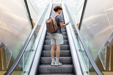 Havaalanında yürüyen merdivende elinde akıllı telefon ve sırt çantasıyla duran genç bir yolcunun arka görüntüsü. Modern ulaşım ve seyahat konsepti, tam boy