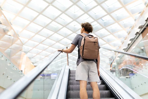 Sırt çantalı gezgin yürüyen merdivende duruyor, tırabzanları tutuyor ve yukarı bakıyor. Parlak tavan ve modern seyahat atmosferi, boşluğu kopyala