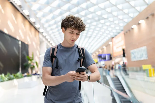 Havaalanı terminalindeki genç adam akıllı telefon kullanıyor, yürüyen merdivenin yanında duruyor. Gezgin adam cep telefonundan mesajları ya da uçuş detaylarını kontrol ediyor.