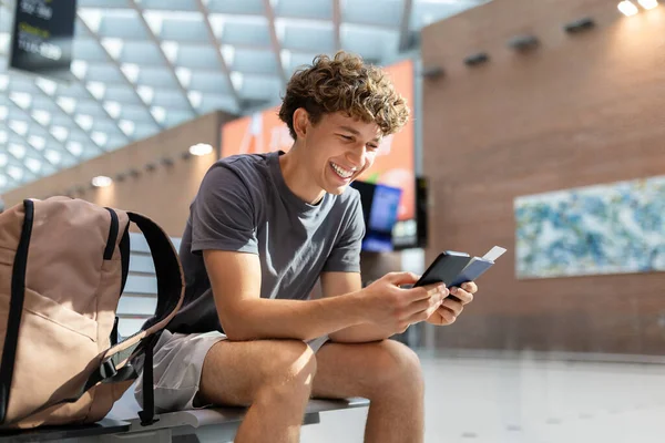 Havaalanı bankında oturan, elinde akıllı telefon ve pasaportla gülen genç adam. Neşeli gezgin biniş detaylarını kontrol ediyor, kopyalama alanı